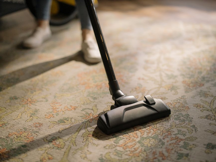 A person kneeling on a patterned rug in a residential room, handling a yellow and black portable vacuum cleaner with a flexible hose. The individual is wearing a light-colored jacket and blue jeans, and their hands are adjusting the vacuum's lid, revealing its interior. The room features wooden furniture and warm lighting, emphasizing surface cleaning and deep cleaning processes in a domestic setting, aligning with services offered by Carpet Cleaning Lambeth for period homes near Lambeth Palace SE1.
