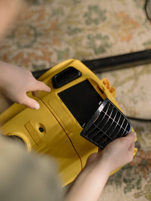 A person kneeling on a patterned area rug, using a yellow vacuum cleaner with a black hose and handle to perform surface cleaning. The individual, wearing a khaki shirt and blue jeans, is opening the vacuum’s top compartment, revealing dust collection elements inside. The area rug has intricate floral patterns in shades of beige, green, and red, and is situated on a wooden floor. The lighting in the room is soft and warm, emphasizing the cleanliness of the carpet and the equipment's operational condition. The scene illustrates domestic cleaning activity aligned with deep cleaning and sanitisation practices by Carpet Cleaning Lambeth for end of tenancy cleaning in Vauxhall SE11.