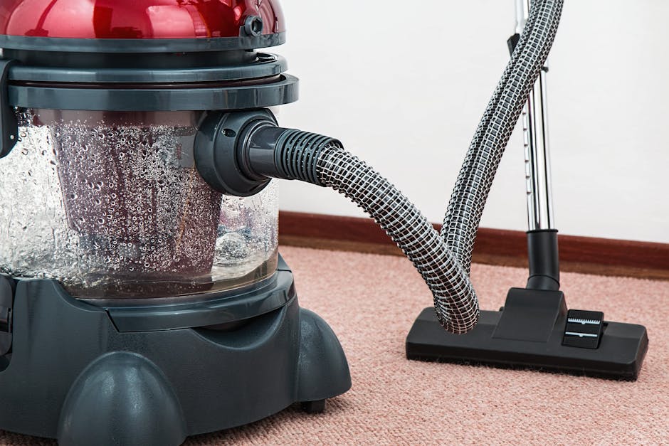 Close-up of a professional vacuum cleaner with a red and black body, positioned on a plush, pinkish-beige carpet in a residential setting. The vacuum's transparent dust container shows collected debris and moisture, indicating recent deep surface cleaning. Attached is a flexible, black-and-white patterned hose connected to a black cleaning head, which rests on the carpet as part of the cleaning process. The background features a simple white wall with a brown wooden skirting board, suggesting tidy interior decoration. The scene highlights equipment used for domestic carpet cleaning and surface sanitisation, emphasizing cleanliness and maintenance facilitated by Carpet Cleaning Lambeth.