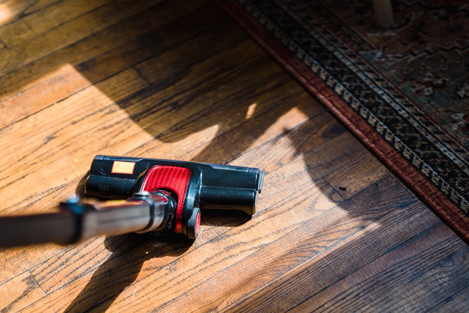 A handheld vacuum cleaner with a black and red body is placed on a wooden floor adjacent to a patterned area rug. The wooden surface shows natural grain and a slightly glossy finish, reflecting light from the left side of the image. The vacuum cleaner’s nozzle is directed toward the floor, indicating surface cleaning activity, while the surrounding area appears clean and dust-free. The area rug features intricate patterns with shades of dark red, beige, and blue, and is positioned in the top right corner of the image, partially covering the wooden flooring. The scene is well-lit, highlighting the smooth texture of the wood and the details of the cleaning equipment, representing professional domestic cleaning services offered by Carpet Cleaning Lambeth, as referenced on the page for rug cleaning in Streatham Hill SW16.