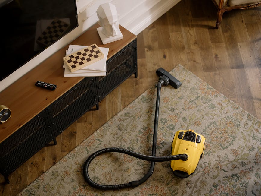 Close-up of a professional vacuum cleaner with a red and black body, positioned on a plush, pinkish-beige carpet in a residential setting. The vacuum's transparent dust container shows collected debris and moisture, indicating recent deep surface cleaning. Attached is a flexible, black-and-white patterned hose connected to a black cleaning head, which rests on the carpet as part of the cleaning process. The background features a simple white wall with a brown wooden skirting board, suggesting tidy interior decoration. The scene highlights equipment used for domestic carpet cleaning and surface sanitisation, emphasizing cleanliness and maintenance facilitated by Carpet Cleaning Lambeth.