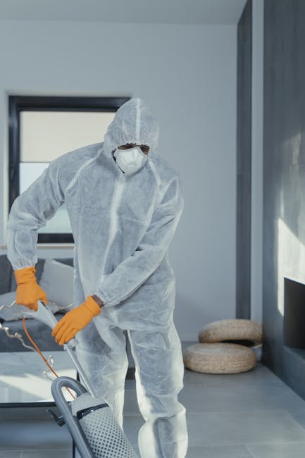 A professional cleaning technician dressed in a full-body white protective suit, face mask, goggles, and orange gloves, is engaged in deep cleaning a modern indoor space. The individual is using a portable cleaning device, possibly a steam cleaner or sanitisation tool, on a smooth tiled floor in a well-lit room with large windows allowing natural light to illuminate the area. The room features neutral-colored walls, a comfortable cushioned seat, and a sleek, minimalistic design. Visible surfaces include polished tiles and glass windows, and the overall space appears clean and well-maintained. This setting exemplifies thorough surface cleaning and sanitisation practices, aligning with the services provided by Carpet Cleaning Lambeth, as described on their website for same-day deep cleaning solutions, addressing common delays and issues related to maintaining hygiene in residential and commercial environments.
