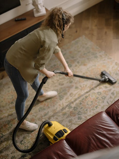 A woman is engaged in surface cleaning within a domestic setting, using a yellow vacuum cleaner with a black hose attachment on a patterned fiber rug. She is dressed in casual attire, including a beige blouse and dark jeans, and is bending slightly forward to handle the vacuum cleaner's hose and nozzle. The room features a wooden flooring section visible near the rug, a dark brown leather sofa in the foreground, and a sideboard with decorative items and a window providing natural light. The space appears clean and well-maintained, reflecting professional deep cleaning processes associated with Carpet Cleaning Lambeth's services in Brixton Market SW9.