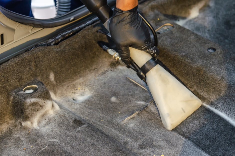 A professional technician, wearing black gloves, is performing surface cleaning on a grey carpeted floor in a commercial or residential setting. The technician is holding a white spray nozzle connecte