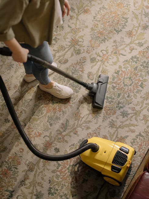 A professional technician, wearing black gloves, is performing surface cleaning on a grey carpeted floor in a commercial or residential setting. The technician is holding a white spray nozzle connecte